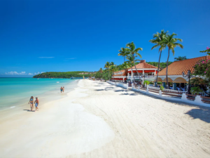 The beach at Sandals Grande Antigua with the ocean to the left and the resort to the right with people walking in the sand and palm trees