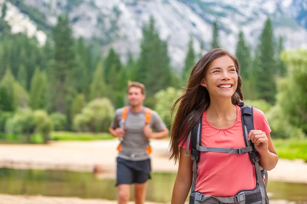 Hikers walking in nature forest of Yosemite. Happy campers hiking through trail trek in mountains. Asian girl with backpack looking up, man friend behind.